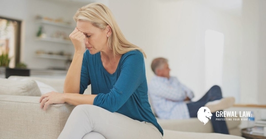 Woman sitting on a couch with her hand on her head and a man in the far background with his arms and legs crossed.